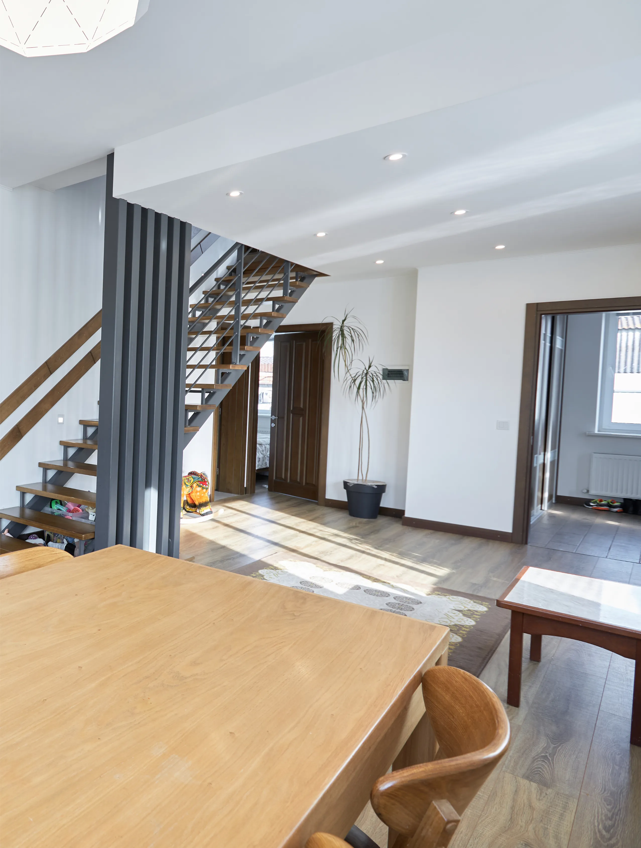 A modern, open-concept living space with a wooden table, chairs, and a metal staircase. The room is lit by ceiling lights and natural sunlight from large windows.