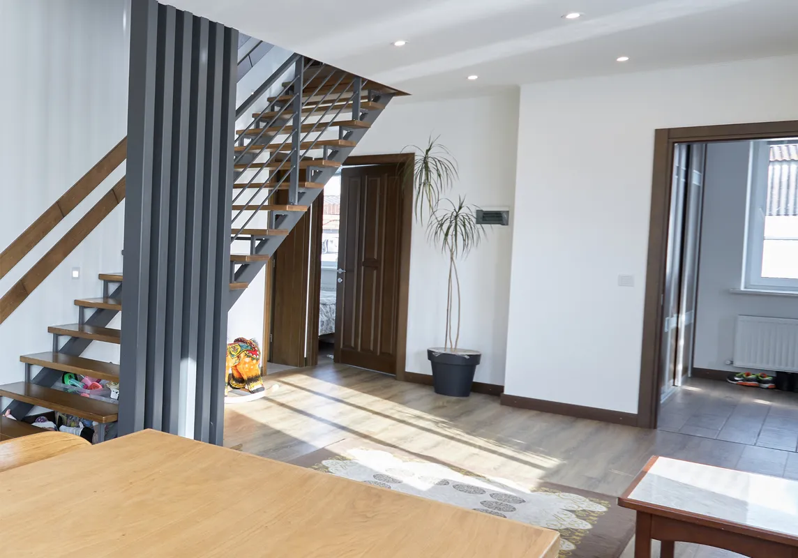 A modern, open-concept living space with a wooden table, chairs, and a metal staircase. The room is lit by ceiling lights and natural sunlight from large windows.