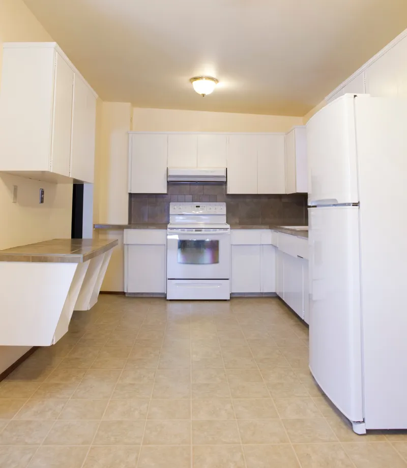An older kitchen with white cabinets, beige tile flooring, and white appliances.